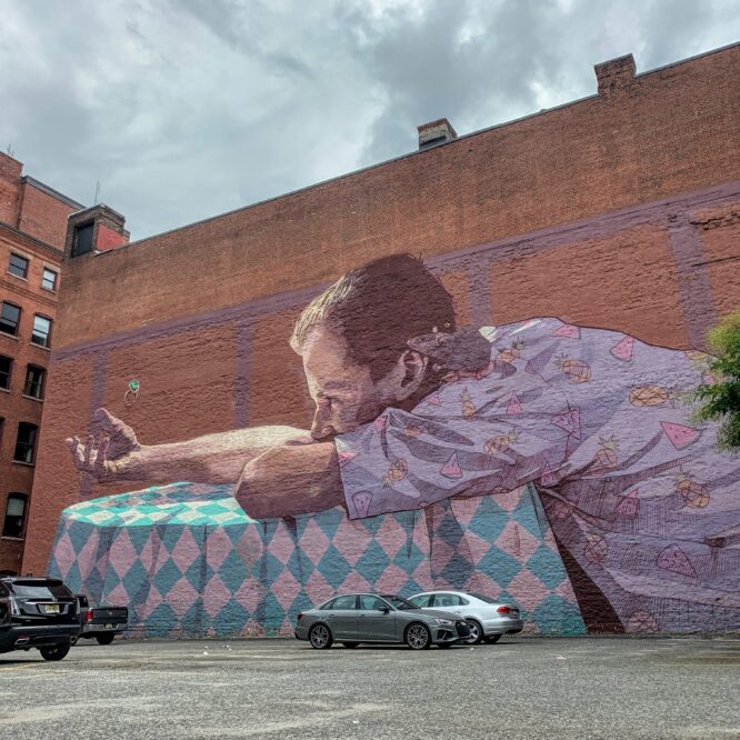 A mural of a child laying forward on a table looking at a wing covers the entire side of a brick building.